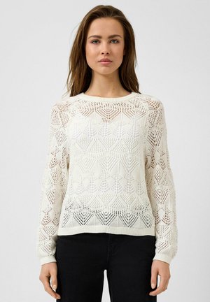 Jonge vrouw die een witte gebreide trui met geometrische patronen en een zwarte broek draagt, staand tegen een effen lichte achtergrond.