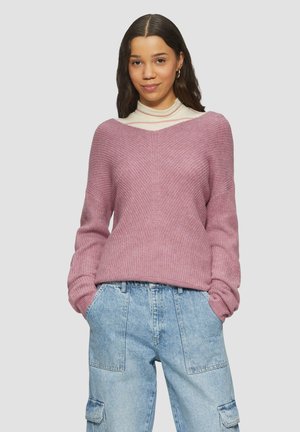 V-Ausschnitt, langärmeliger Pullover aus hellrosa, strukturiertem Strickmaterial, kombiniert mit hochgeschnittenen blauen Denim-Cargohosen mit großen Taschen.