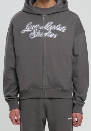 Osoba nosząca ciemnoszarą bluzę z kapturem i zamkiem błyskawicznym z napisem „Low Lights Studios” oraz pasujące spodnie z małym logo „lowlights” na lewej nodze.