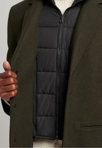 Manteau extérieur vert olive avec une doublure matelassée noire, doté d'une fermeture zippée et d'une texture matelassée, porté sur un pull crème.