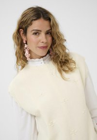 Kermavaatteinen neuleliivi, jossa on pyöreä pääntie, kukkakuosiset tekstuurit ja ribbaushelma, kerrostettuna valkoisen kauluspuseron päälle.