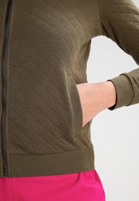 Vadderad olivgrön jacka med dragkedja och ribbade ärmslut, som har en framficka med horisontell öppning.