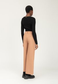 Person står med ryggen til, iført en tætsiddende sort langærmet top, beige vidde benbukser, sorte strømper og klodsede sorte sko.
