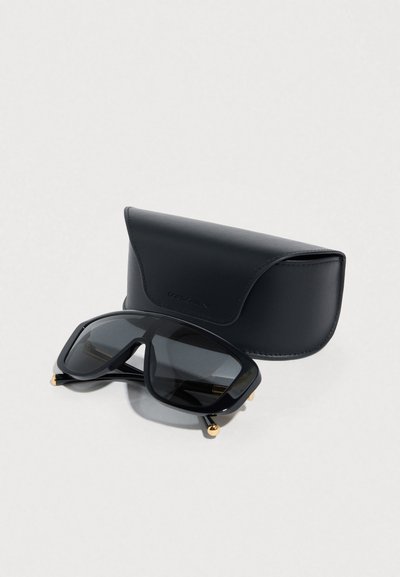 Lunettes de soleil noires surdimensionnées avec une monture géométrique audacieuse, des verres teintés gris, des accents dorés aux branches, et un étui en cuir noir.