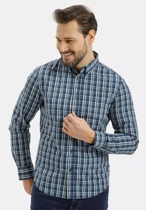 Mężczyzna uśmiechający się, ubrany w niebiesko-białą kraciastą koszulę na guziki z długimi rękawami, stojący na jednolitym jasnym tle.