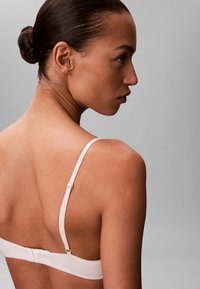 Reggiseno rosa chiaro con spalline regolabili e chiusura a gancio. Tessuto liscio, design minimale e una leggera lucentezza. Vista laterale.