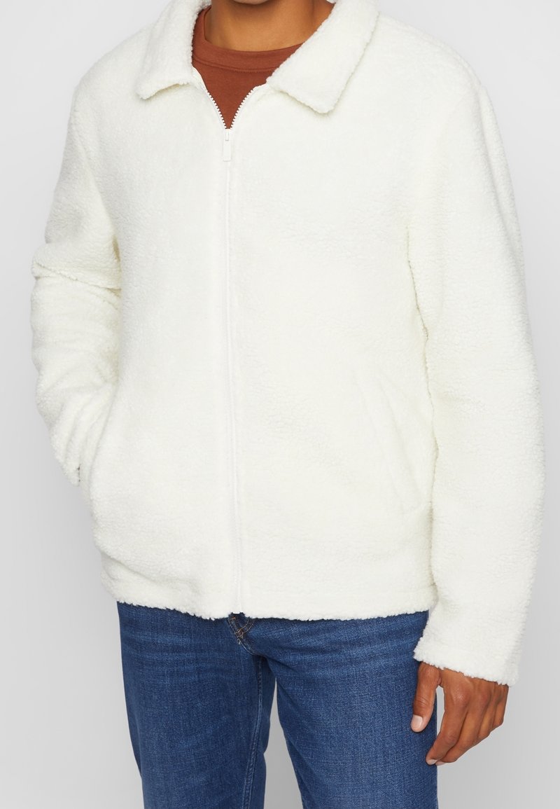 White faux shearling zip-up jacket with a collar, long sleeves, and elastic cuffs, paired with blue denim jeans. Soft texture, casual design.