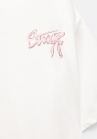 T-shirt blanc en coton avec un logo graphique rose et un nœud sur le côté gauche de la poitrine. Coupe simple et décontractée avec des manches courtes.