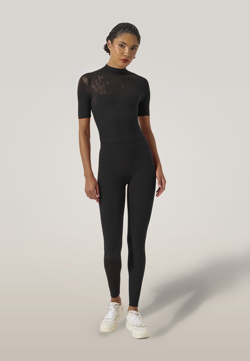 Svart figurnära bodysuit med korta ärmar och hög krage. Har ett genomskinligt blommönster av spets på en axel. Bärs med vita sneakers.