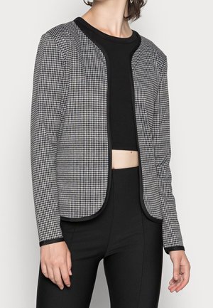Femme portant une veste ouverte à motifs noir et blanc sur un top court noir et un pantalon noir taille haute.