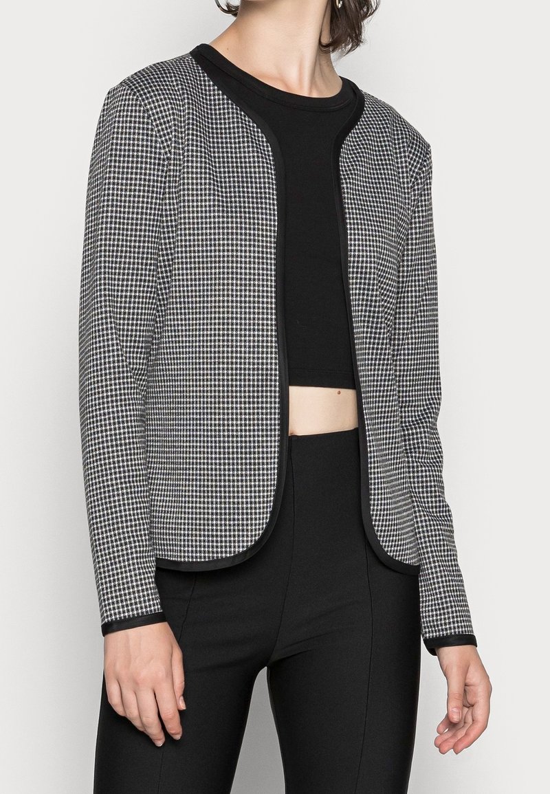 Femme portant une veste ouverte à motifs noir et blanc sur un top court noir et un pantalon noir taille haute.