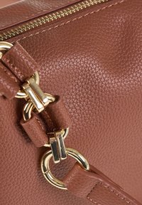 Dettaglio di una borsa in pelle marrone con texture, anelli in metallo dorato e una cerniera, che mette in risalto il fissaggio della tracolla e le cuciture.