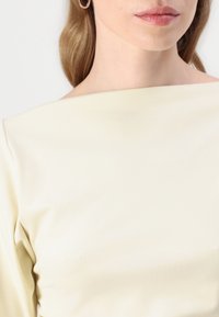 Haut crème jaune dégageant les épaules avec une texture lisse et mate. Gros plan sur le décolleté et la poitrine, mettant en avant la simplicité du design.