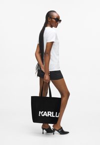 Fekete bevásárlótáska nagy fehér "KARLLA" felirattal, fehér pólóval és fekete sorttal párosítva. A modellen fekete napszemüveg és magassarkú mules szandál van.