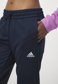 Námornícke športové nohavice s elastickým pásom, s jemným leskom, bočnými vreckami a bielym vyšitým logom Adidas na ľavom stehne.