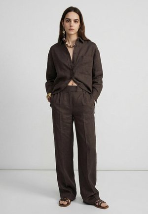 Femme debout vêtue d'une chemise ample en lin brun foncé et d'un pantalon large assorti, accessoirisée avec des colliers, des boucles d'oreilles, des bracelets et des sandales.
