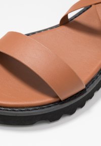 Ruskealla synteettisellä sandaalilla on kaksi leveää ristikkäistä hihnaa, teksturoitu pinta ja paksu musta kumipohja, jossa on urakuvio.