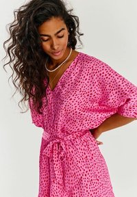 Růžové šaty s výstřihem do V, krátkými rukávy a zavazovací šňůrkou v pase. Materiál má hladkou texturu a je pokrytý rozptýlenými červenými tečkami.