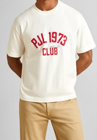 Krémové bavlněné tričko s krátkými rukávy, na přední straně s červeným a modrým textem "PJL 1973 CLUB PHYS. ED.". Měkká textura, uvolněný střih.