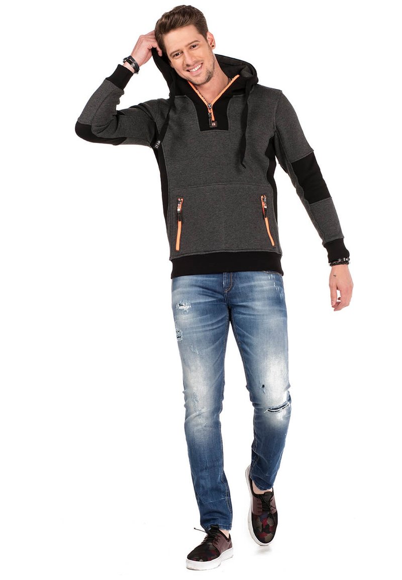 Grijze en zwarte capuchontrui met oranje ritsen en twee zijzakken, gecombineerd met versleten blauwe jeans en gepatternde schoenen.