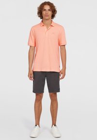 Polo shirt corail à manches courtes avec une texture subtile, associé à un short gris foncé. Des baskets blanches complètent la tenue.