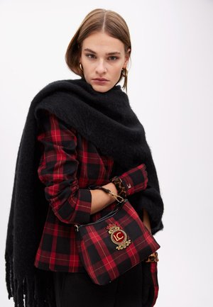 Woman wearing a red and black plaid jacket and matching bag, with a large black scarf draped over her shoulders on a white background.