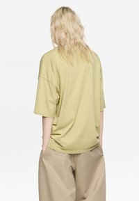 Camiseta de algodón de corte holgado y manga corta en verde pálido, con hombros caídos y dobladillo redondeado. Combina con pantalones de pierna ancha beige.