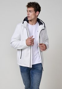 Jachetă albă tip softshell cu glugă, căptușeală interioară neagră, două buzunare cu fermoar și fermoare contrastante. Modelul poartă un tricou alb și blugi blue.