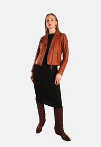 Kvinde iført en brun læderjakke, sort midi-kjole og brune knæhøje støvler, stående imod en ensfarvet lys baggrund.