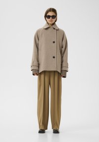 Beige wollen oversized jas met een kraag, knopen aan de voorkant en brede manchetten. Gepaard met hooggeritste, geplooide bruine broek.