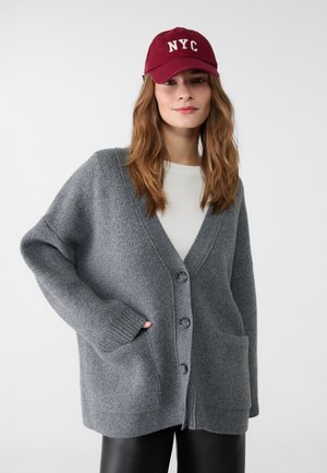 Grå strikket cardigan med V-udskæring, knaplukning og forlommer. Kombineret med en vinrød kasket med "NYC" i hvid skrift.