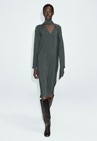 Lungo vestito grigio ardesia con scollatura a V e dettaglio di sciarpa drappeggiata. Presenta maniche lunghe e una silhouette dritta. Indossato con stivali aderenti al ginocchio.