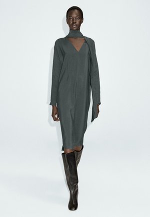 Lang, leisteengrijze jurk met een V-hals en een gedrapeerde sjaal detail. Heeft lange mouwen en een rechte silhoeet. G gedragen met strakke, kniehoge laarzen.
