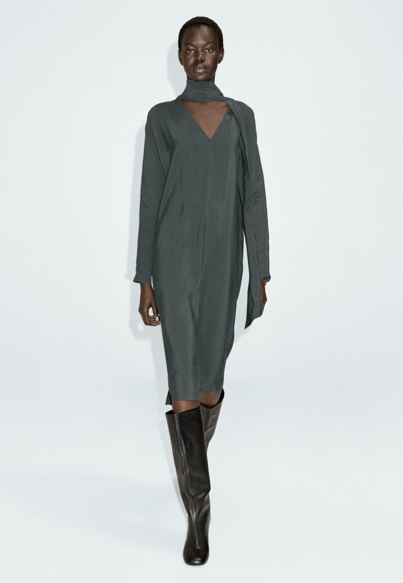 Długa, szaro-skalista sukienka z dekoltem w serek i drapowaną detalem w postaci szalika. Posiada długie rękawy i prosty krój. Noszona z dopasowanymi, wysokimi na kolana botkami.