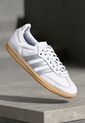 Hvid Adidas Samba sneaker med gummisål, sølvstriber og snørebånd, vinklet over en betonoverflade mod en mørk baggrund.