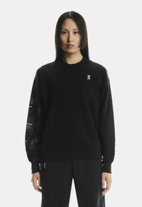 Fekete pulóver hosszú ujjakkal, fehér logóval a mellrészen és textúrázott mintázattal a bal ujján. Puha anyag, laza fazon.