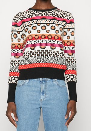 Person wearing colorful patterned sweater with animal print, sun motifs, stripes, and geometric designs paired with light blue jeans.