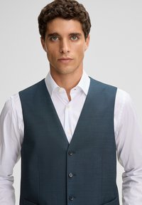 Młody mężczyzna z kręconymi brązowymi włosami, ubrany w ciemnoniebieską kamizelkę na białą koszulę z guzikami, patrzy prosto przed siebie z neutralnym wyrazem twarzy.