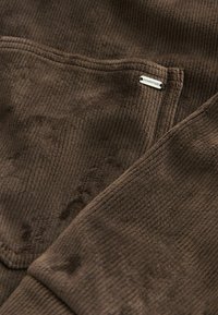 Ruskea corduroy-kangas, jossa on pehmeä rakenne ja näkyvä ribbitetyyli. Siinä on pieni metallilappu, jossa lukee "DOWNTIME".