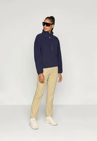 Granatowy polar z pół zamkiem i wysokim kołnierzem, połączony z beżowymi spodniami slim-fit i białymi butami sportowymi, noszony z czarnymi okularami przeciwsłonecznymi.