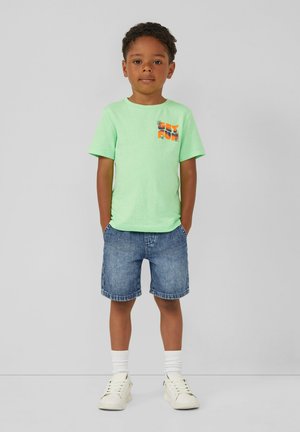Groene katoenen T-shirt met grafische print op de zak, gecombineerd met blauwe denim shorts, witte sokken en witte sneakers. Kind staat met handen in de zakken.