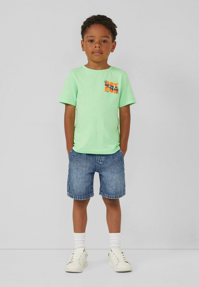 T-shirt verde in cotone con taschino stampato, abbinato a pantaloni corti in denim blu, calzini bianchi e sneaker bianchi. Bambino in piedi con le mani nelle tasche.
