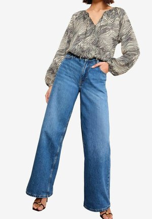 Kobieta ubrana w szerokie, niebieskie dżinsy, koszulę w cętki zebry z długim rękawem oraz czarne sandały na obcasie z odkrytymi palcami, stojąca z jedną ręką w kieszeni.