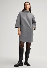 Grijze oversized jurk met een hoge kraag, zijzakken en losse mouwen, gecombineerd met zwarte gemusterde panty's en zwarte kniehoge laarzen.