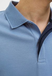 Šviesiai mėlynas polo marškinėliai su tekstūruota, tinklelio tipo apykakle ir tamsiai mėlynu dekoratyviniu kraštu. Su vienu mygtuko užsegimu ir glotnia audinio tekstūra.