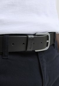 Zwarte leren riem met een zilveren metalen gesp, met een zachte textuur en aanpassingsgaten. Het gemerkte leren band is zichtbaar.