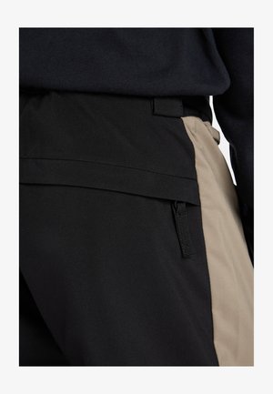 Black and beige dual-tone pants with a smooth texture, featuring a side zip pocket and belt loops; casual design with a modern fit.