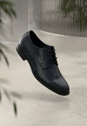 Czarne skórzane formalne buty sznurowane unoszące się nad jasnoszarym popękanym betonowym podłożem, z rozmytym tłem roślin i metalowego ogrodzenia.