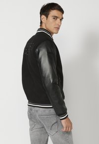 Black varsity jacket with leather sleeves, striped ribbed cuffs, and textured fabric. Features subtle branding on the back.