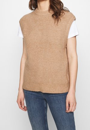 Kobieta w bezrękawniku z dzianiny w kolorze wielbłądzim, nałożonym na białą koszulę z krótkim rękawem i niebieskie dżinsy, stojąca na jednolitym tle.